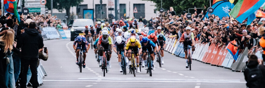 Grupo de ciclistas de carretera en competición, rodeados de espectadores, con un vehículo de apoyo al fondo. Grupo de ciclistas de carretera en competición, rodeados de espectadores, con un vehículo de apoyo al fondo.