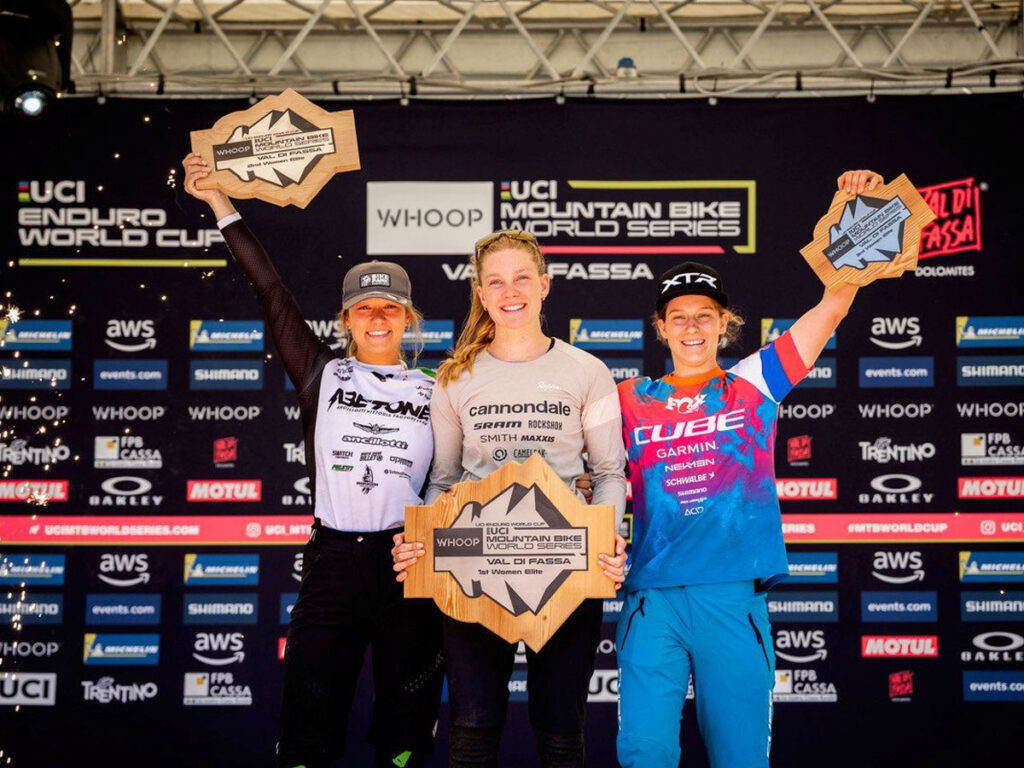 Simona Kuchynkova on the podium of the Enduro World Cup in Canazei, raising her trophy with her left arm, next to the first and second place winners