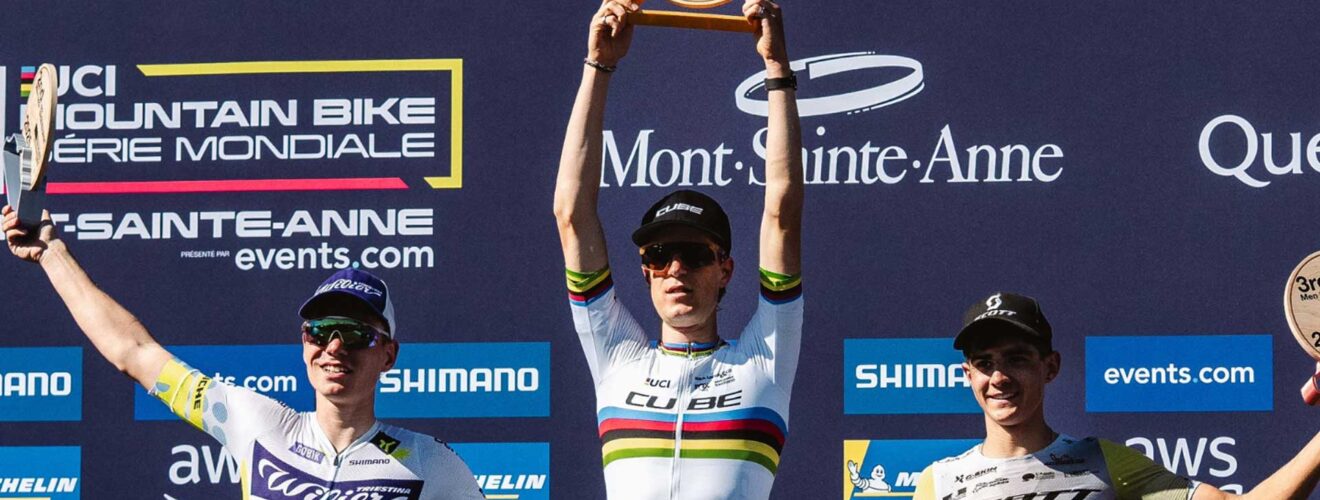 Finn Treudler jubelt beim Cube Factory Racing Saisonfinale 2025 in Mont-Sainte-Anne mit der Trophäe in den Händen, flankiert von dem Zweit- und Drittplatzierten auf dem Podium.